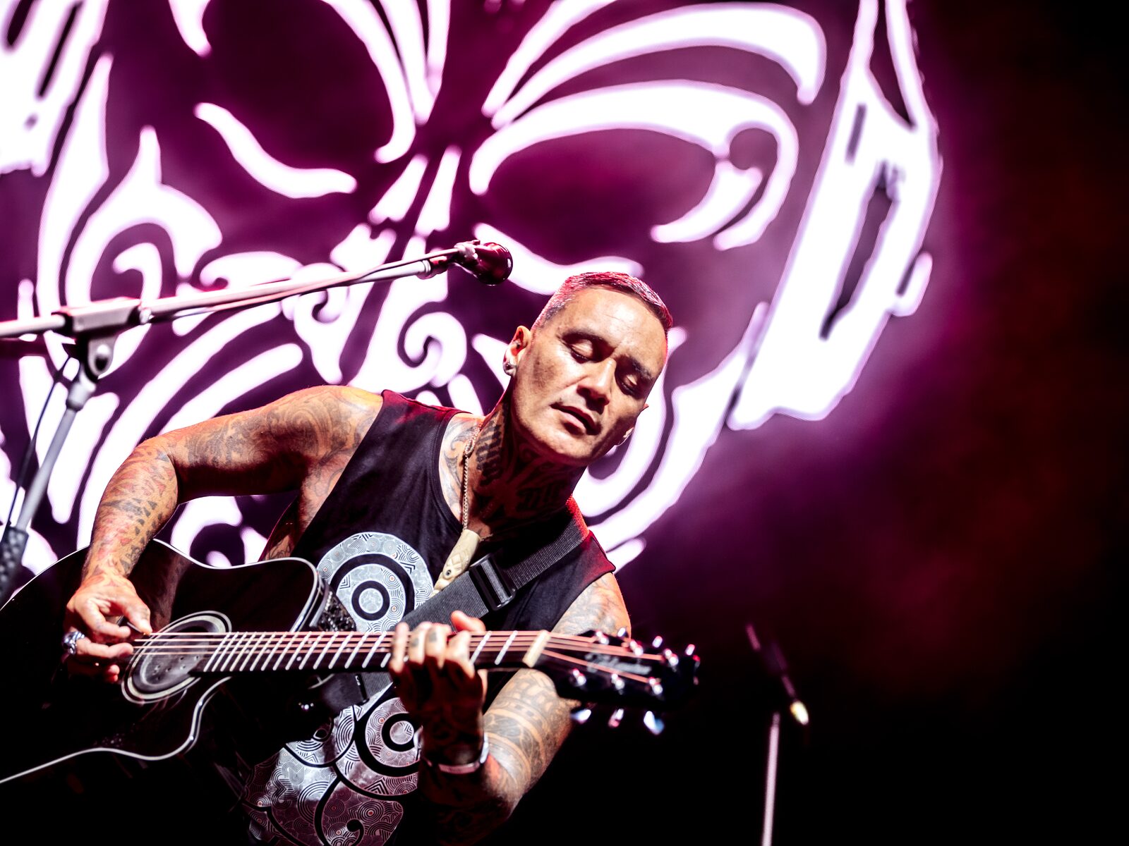 Tiki Taane playing guitar on stage, a moko projection lighting the backdrop